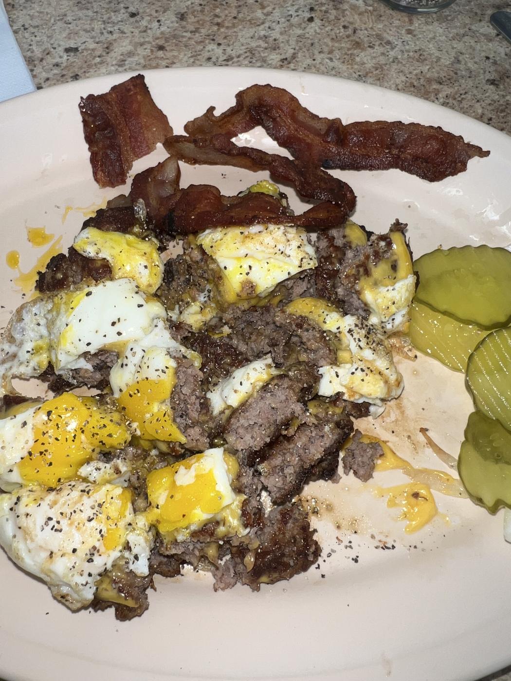A skillet of burger meat, eggs, cheese and bacon with pickle slices on the side